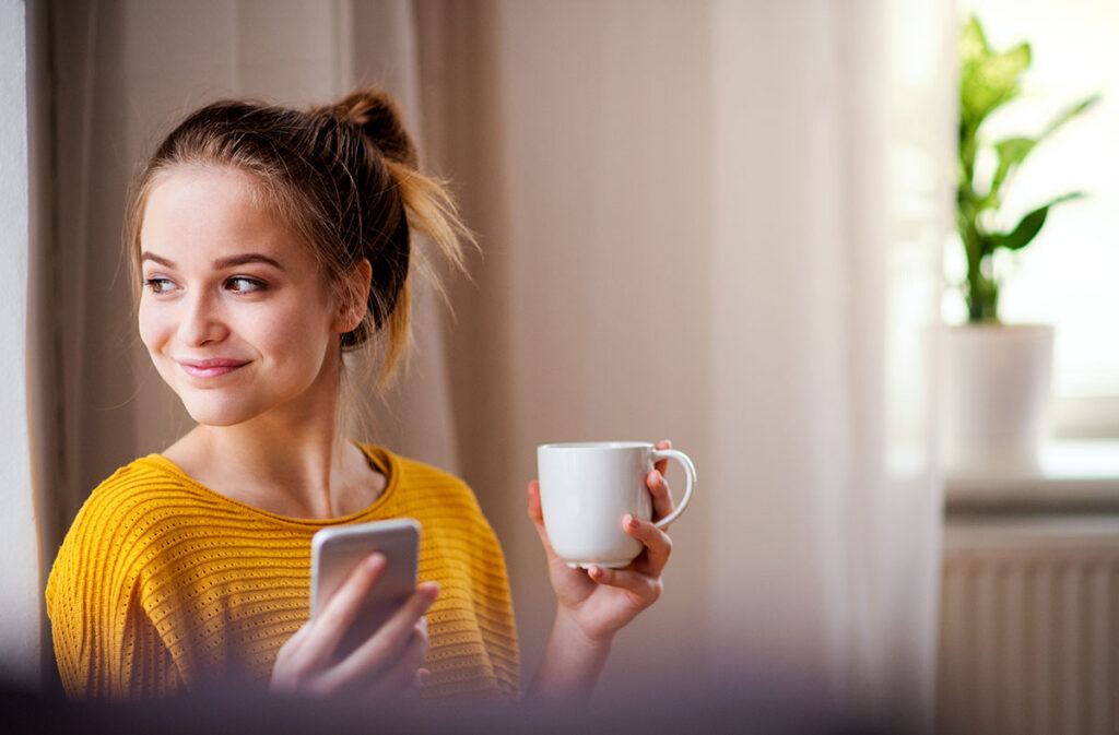 a woman smiles while holding a cup of tea and her phone while thinking about the benefits of cbt