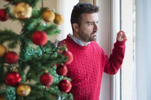 a man standing next to a christmas tree looks out a window wondering how mental health and the holidays are intertwined