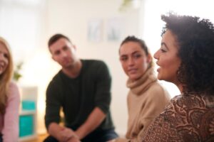 a woman talks in group therapy about being an enabler in addiction and the group of people face and listen to her