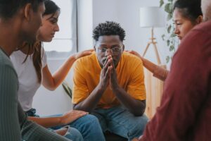 a man sits appearing distraught with his hands near his mouth and two people consoling him while he thinks about his addiction therapy options