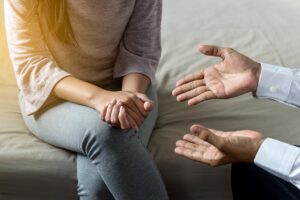 a woman sits on a couch with her hands clasped to each other while her friend discusses the benefits of individual therapy program