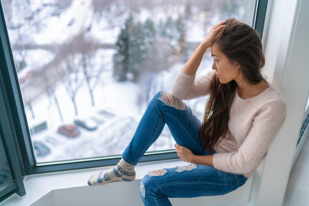 a woman sits on a window sill appearing distraught and thinking about the common causes of depression