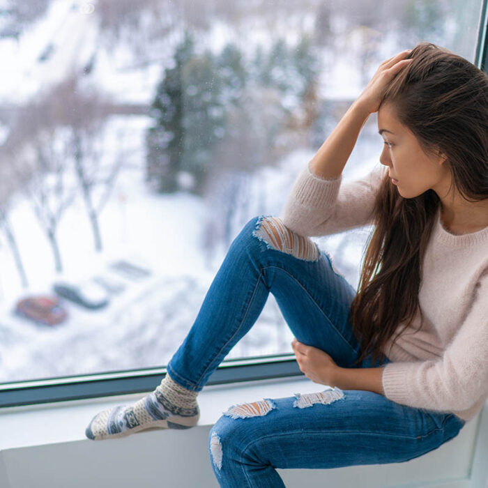 a woman sits on a window sill appearing distraught and thinking about the common causes of depression