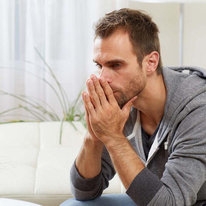 man in hoodie sits on his couch with his hands to his mouth looking worried about the signs of painkiller abuse