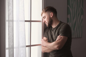 a man with his arms folded and leaning on a wall near a window looks out that window and thinks about finding depression treatment in massachusetts