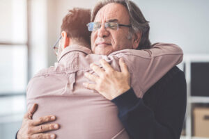 an older man with glasses hugs a younger adult male in their alcohol rehab program