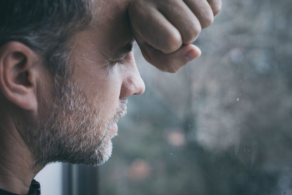 a distraught man leans on a window while looking out the window and thinking about the signs of prescription drug abuse