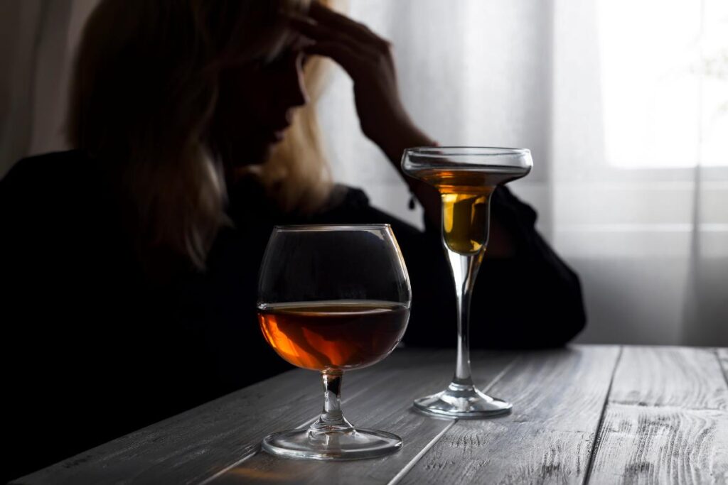 a person sits in the dark leaning on a table with two alcohol beverages in front of them while wondering if they might have a drinking problem