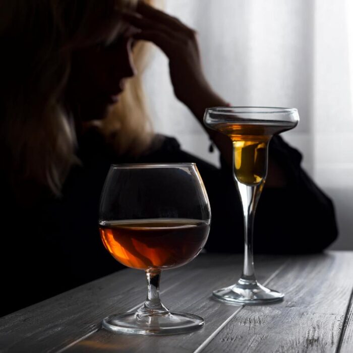 a person sits in the dark leaning on a table with two alcohol beverages in front of them while wondering if they might have a drinking problem