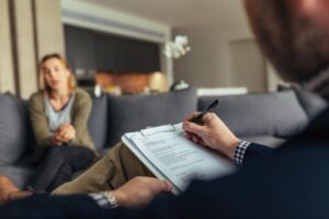 a male therapist sits and takes notes on his clipboard about the woman patient sitting across from him while he explains why mental health care is important