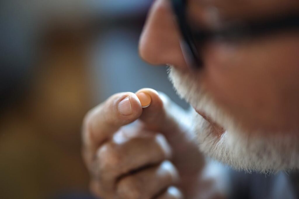 a older man with a beard holds a pill near his mouth after popping pills not prescribed to him