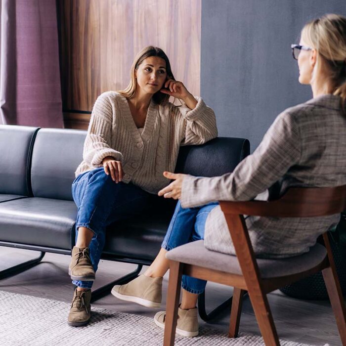 an adult woman sits on a couch while her therapist sits in a chair across from her talking about a partial hospitalization program for addiction